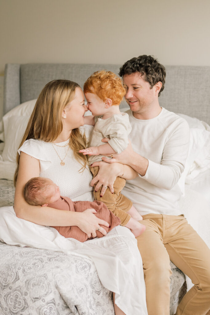 Family with toddler and baby sitting on the bed in neutral colors sharing kisses and love |  Newborn Photos with Toddler with Janet Winter Photography