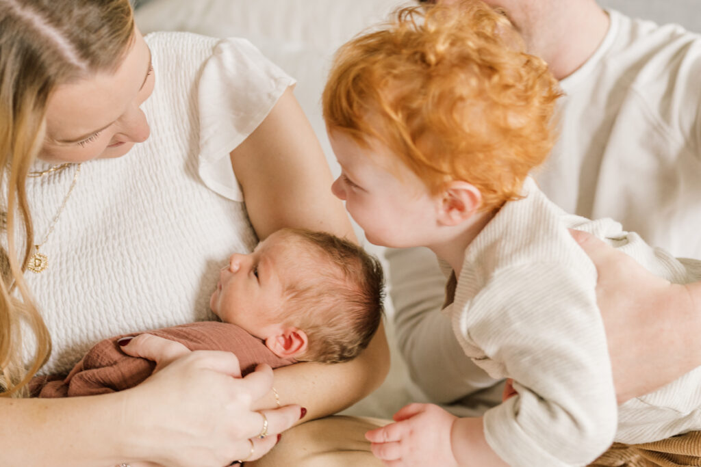 Family with toddler and baby on bed and toddler trying to kiss baby | Newborn Photos with Toddler with Janet Winter Photography 