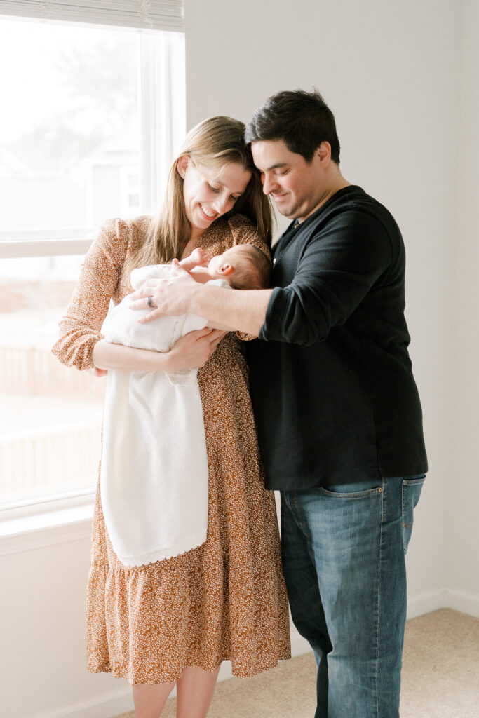 mom and dad holding baby girl near window during newborn photos at home